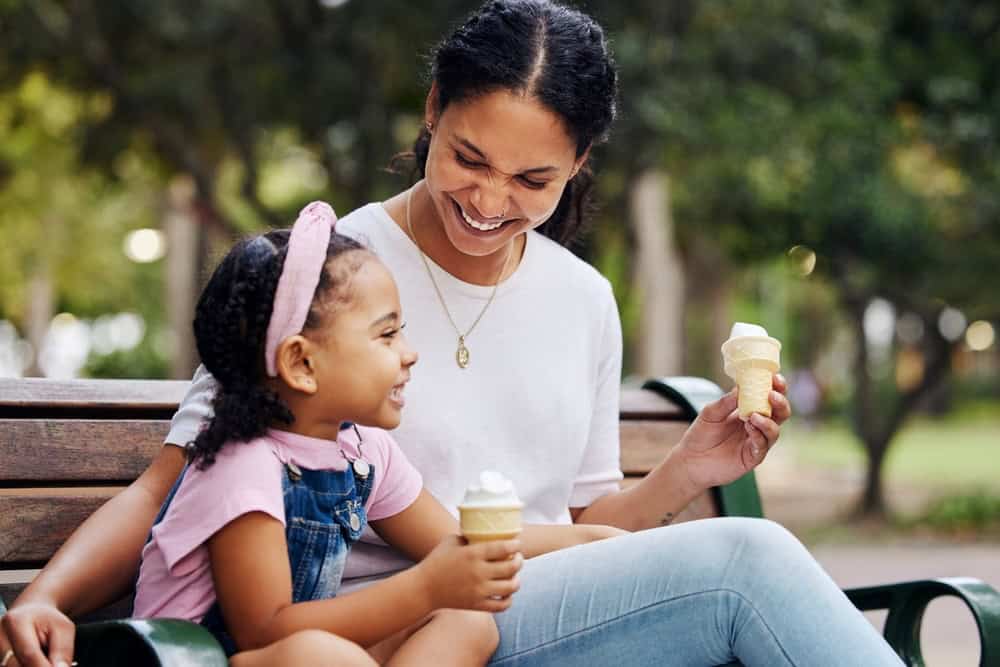 Summer, park and ice cream with a mother and daughter bonding together while sitting on a bench outdoor in nature. Black family, children and garden with a woman and girl enjoying a sweet snack